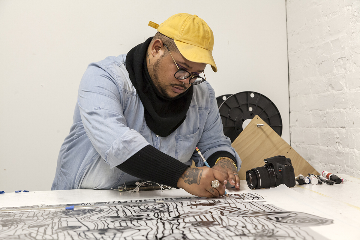 The artist Jonathan Lyndon Chase draws with a pencil to produce their limited artist edition, "Bending $ag." They're wearing a yellow hat and a denim jacket over a warm black under-layer and black scarf.