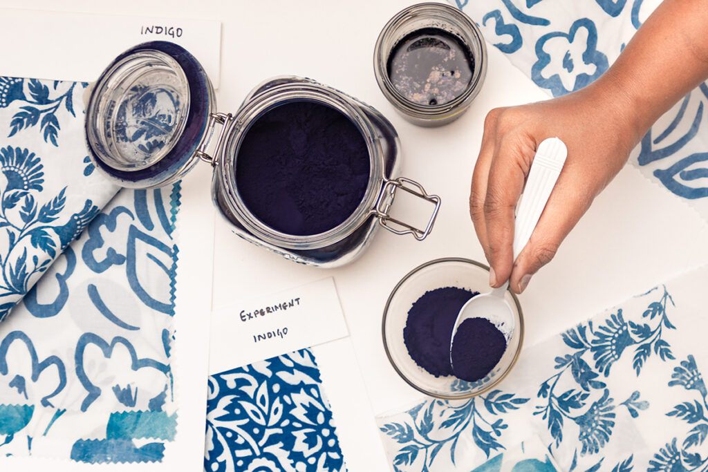 A table spread that includes a jar and cup of indigo with examples of blue printed patterns. A hand reaches over to spoon indigo from the cup.