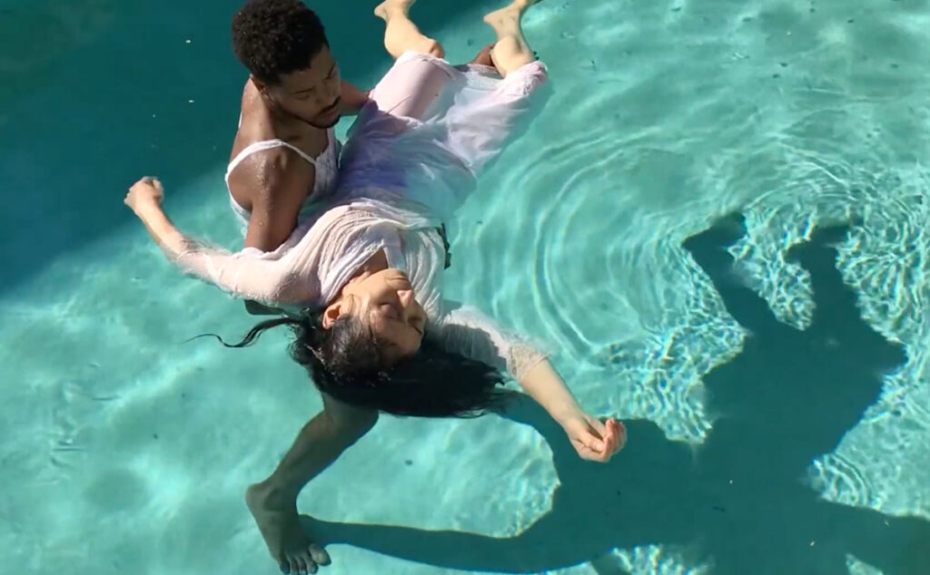 An image of DonChristian Jones, a young Black man, cradling Eiko Otake, an older Japanese woman in a swimming pool. Her arms are outstretched in different directions. Their shadows extend to the right of the image and connect at the man's feet.