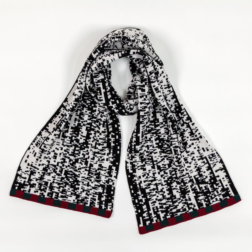 A long scarf with a pixelated black and white pattern wrapped in a way that allows space for someone's neck to fit in the center. The bottom of each end of the scarf features a border of maroon and forest green rectangular shapes.