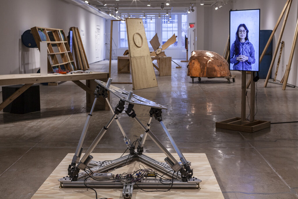 A photograph of a gallery with many disparate wooden and metal sculptures and materials strewn about, some leaning against the walls and others in the middle of the floor. In the foreground is a steel armature with tilted legs and wires. Nearby is a television screen installed sideways on a wooden base. On its screen is a woman with black hair and hands clasped across her body. She is looking at the camera.