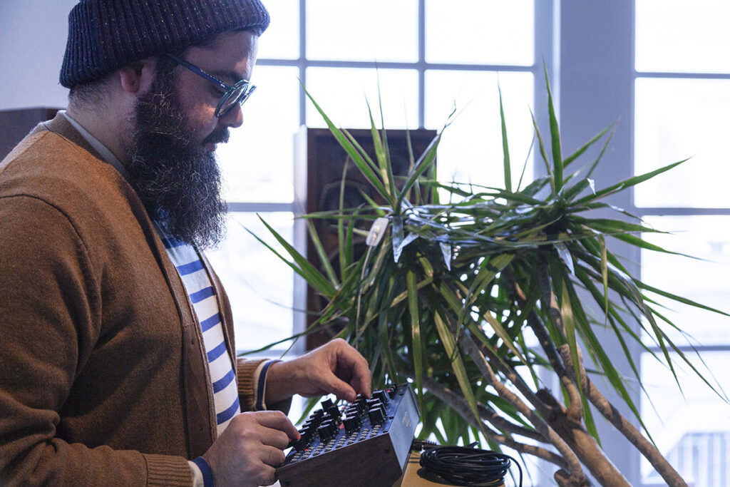 Photograph of artist Raúl Romero, a Puerto Rican-American man with a black bushy beard, black rimmed glasses, a beanie and a brown cardigan, at work near a plant with long spiny leaves. His fingers are on the dials of an electronic device.