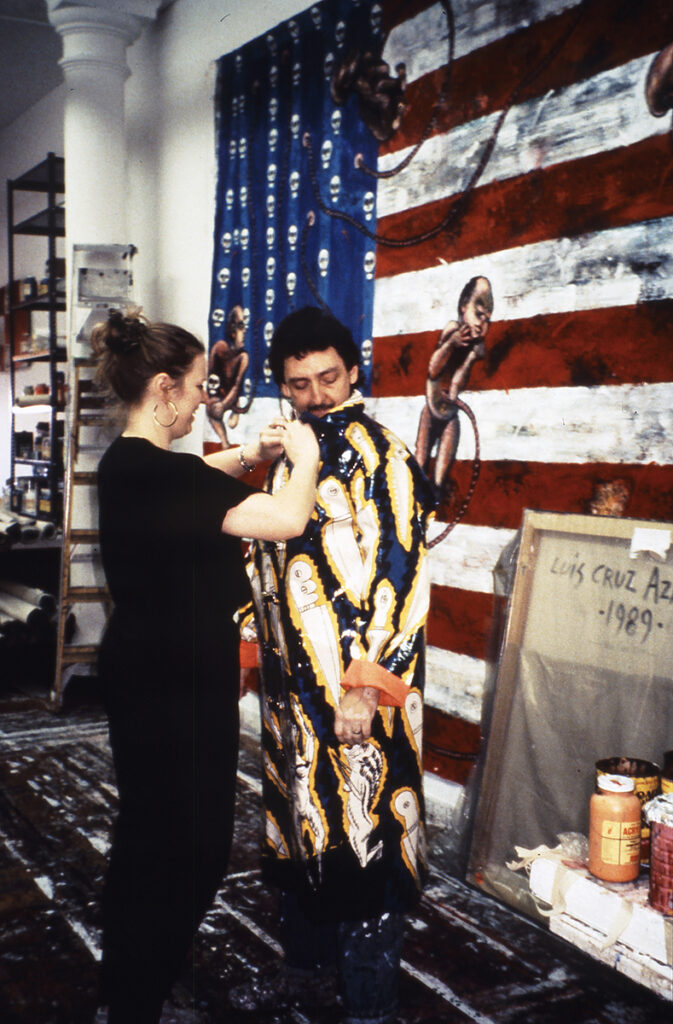 Luis Cruz Azaceta wears his “Acid Rain” coat with the help of his wife, Sharon Jacques, who adjusts his collar while he looks over his left shoulder. Standing in the artist's studio in Soho, New York, the two appear in front of a large painting featuring the American flag with a figure visible just over the artist's left shoulder.