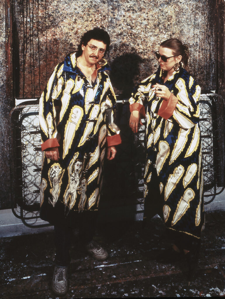 Luis Cruz Azaceta, at left, and his wife, Sharon Jacques, model his “Acid Rain” coat in the artist's studio in Soho, New York. The rain jackets feature cartoonish knives on a background of dark blue. The interior of the jackets, visible on the rolled up sleeves, feature bright orange lining. The two are standing in front of an exposed box spring with a rust colored painting behind it.