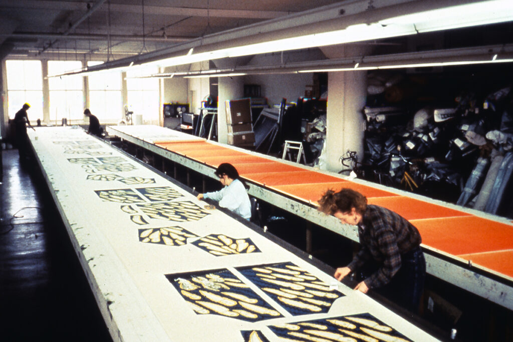 A photograph of a studio featuring long printmaking tables with florescent lights overhead. On the table nearest to the camera, four people work on a repeated pattern of shapes intended for Luis Cruz Azaceta's 