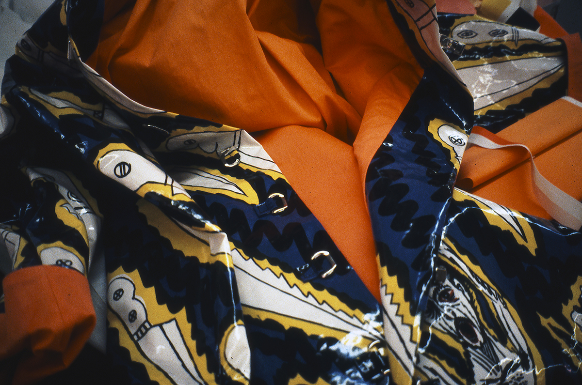 A detail of the collar of a rain jacket with a reflective, plastic sheen features cartoonish knives on a background of dark blue. The interior of the jacket features bright orange lining.
