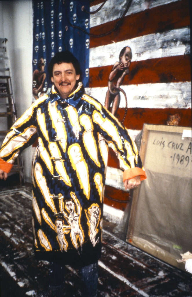 Luis Cruz Azaceta models his “Acid Rain” coat in the artist's studio in Soho, New York. The rain jacket features cartoonish knives on a background of dark blue. The artist gazes at the camera with his arms are spread out. He is standing in front of a large painting featuring an American flag with a figure painted over it, visible just over the artist's left shoulder.