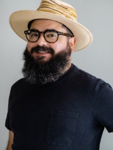 A portrait of Raúl Romero, a Puerto Rican-American man with a bushy black beard. He is wearing a black t-shirt, black rimmed glasses, and a tan sun hat.