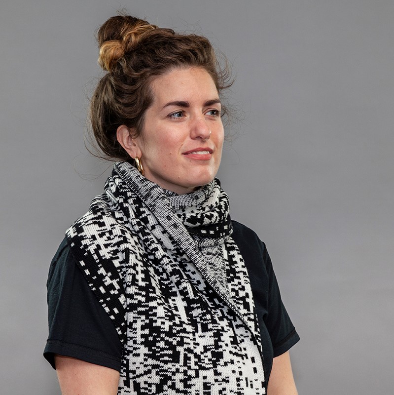 A white woman models a heavy scarf made up of small pixelated shapes of black, grey, and white that give the impression of light on the surface of a body of water.