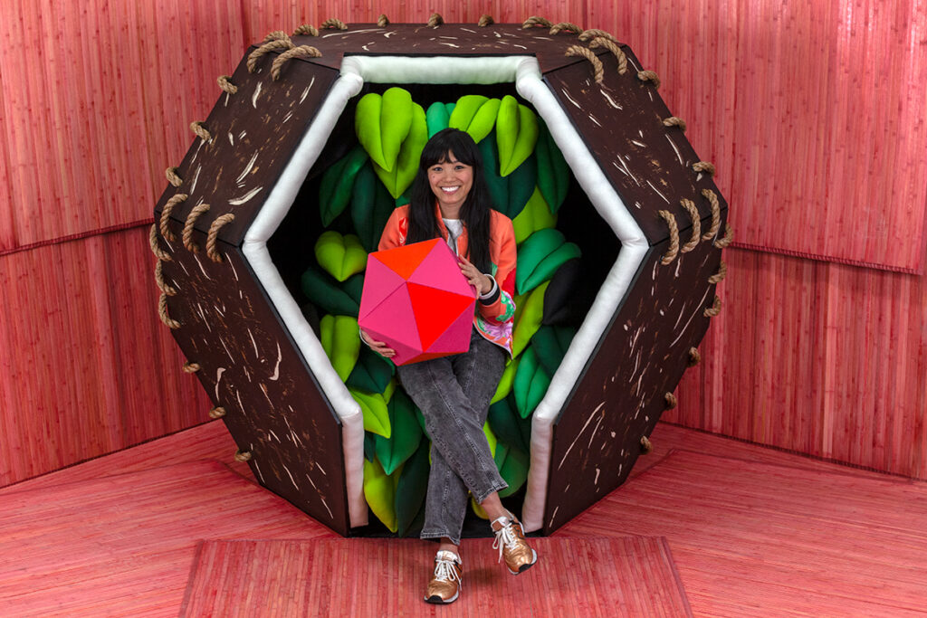 A photograph of the artist Risa Puno, a young woman with black hair, a warm colored blouse and grey jeans, sitting in a brown geometric pod. She is holding a pink and orange multi-sided structure in her hands. Her seat is made of green felted leaves. The walls and floors are all made of the same pink bamboo.
