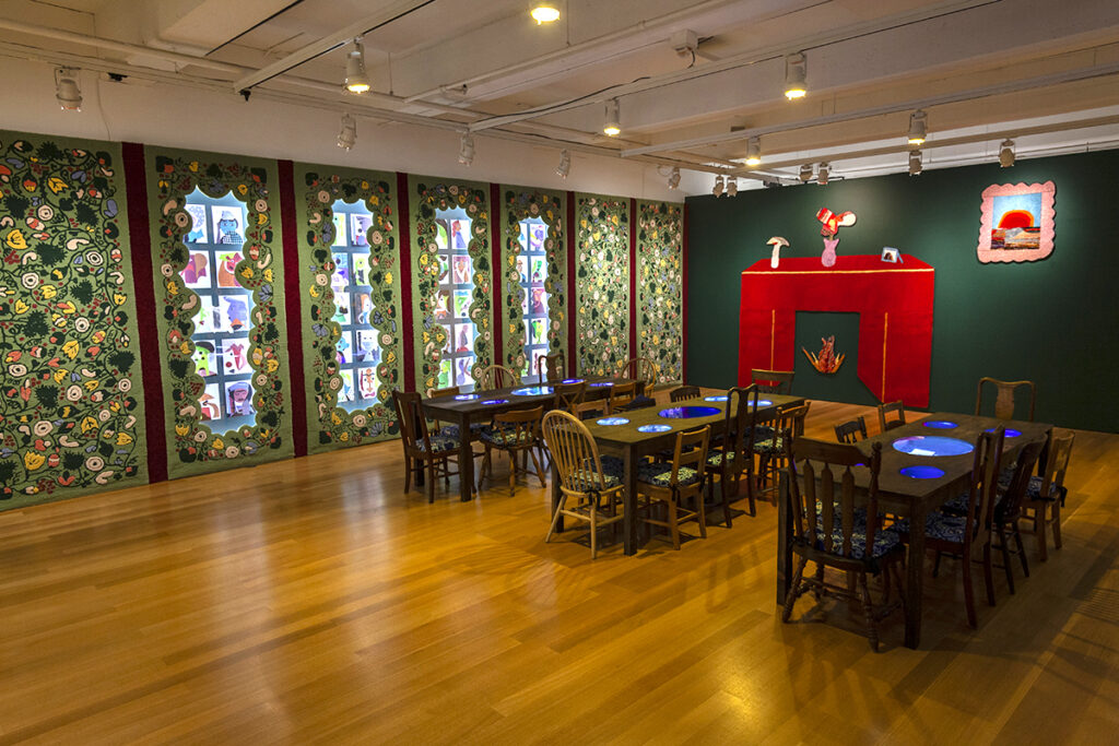 A photograph of a dimly lit room with three wooden tables surrounded by chairs. The far wall is dark green with a large artwork resembling a red fireplace and framed picture. On the left wall is a lighter green tapestry with a busy floral pattern.