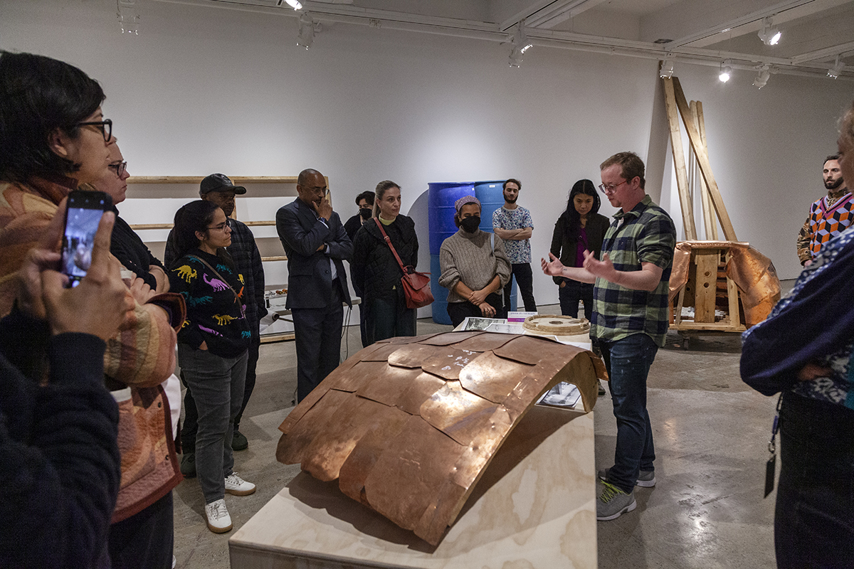 A group of people gather around a large copper sculptural shell in an art gallery. At right, the artist Paper Buck gestures as he describes his artistic practice.