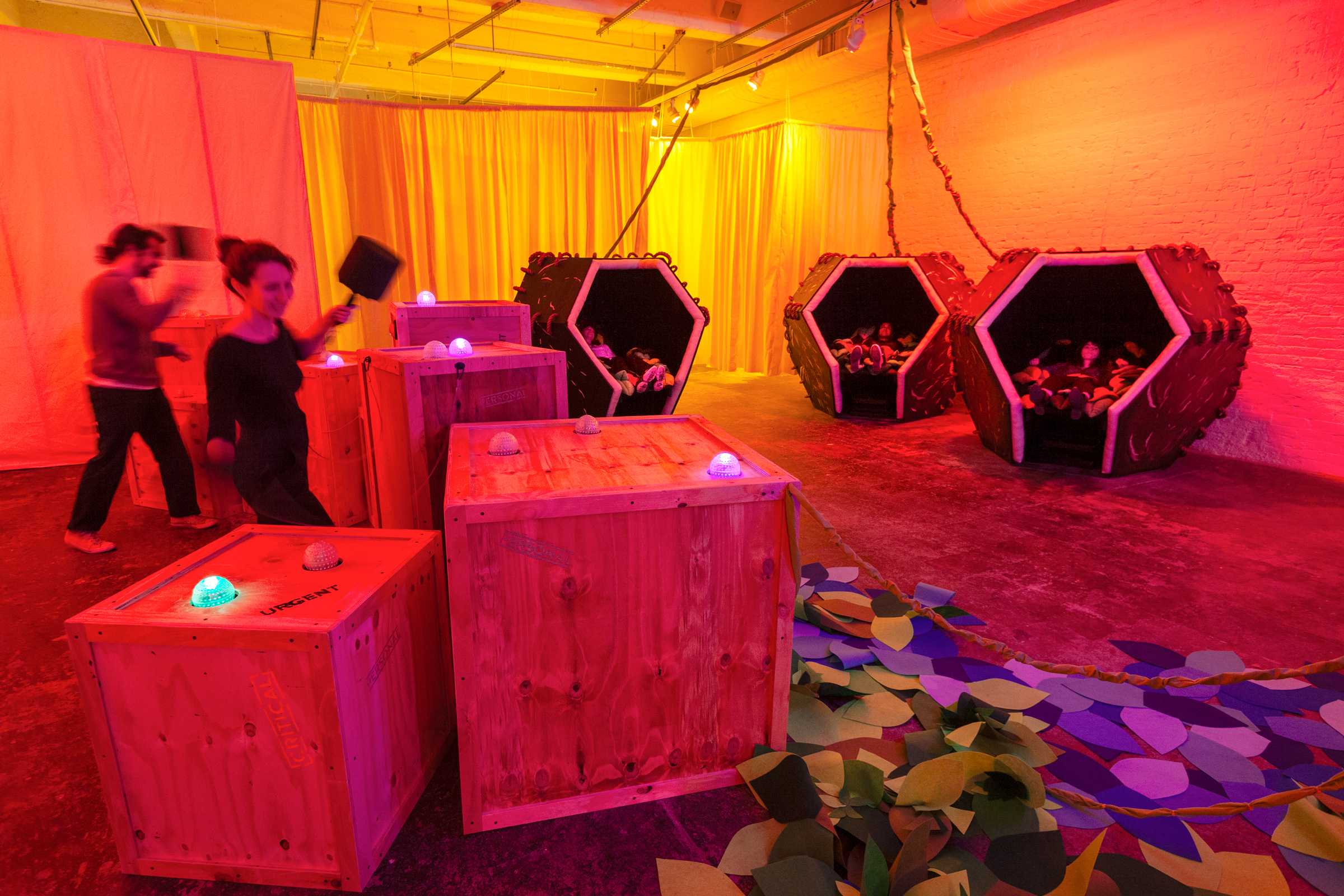 A view of a gallery bathed in pink/orange light. Two people (at left) play a Wac-a-mole-like game with colorful orbs that light up on crates. Three hexagonal-shaped pods sit in the back-right of the room.