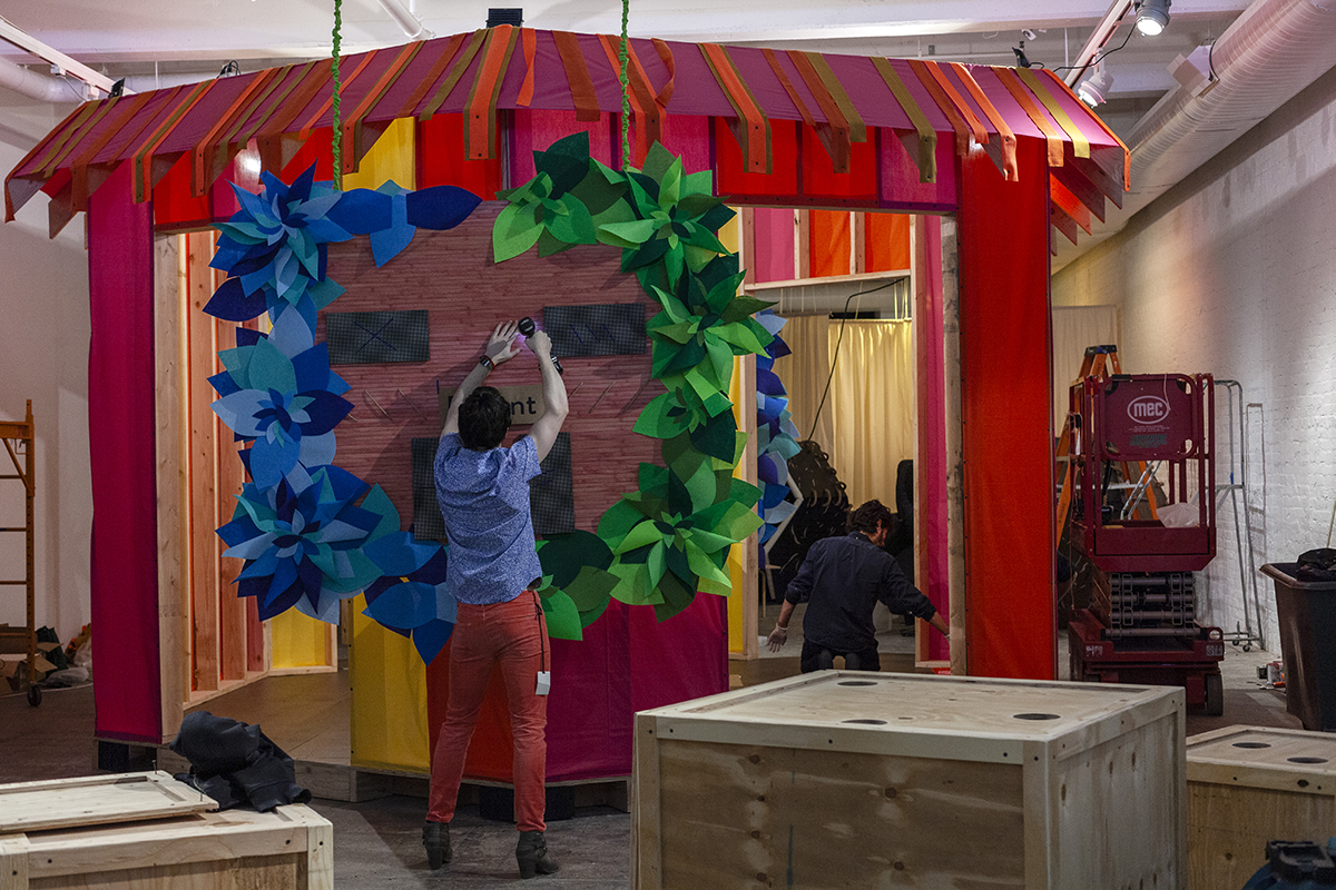 A photograph of a colorful gallery installation with a central structure. At the center, a studio artist works on a scoreboard, displaying green and blue leaves around its perimeter. The larger structure features red, pink, and orange fabric on its roof and walls.