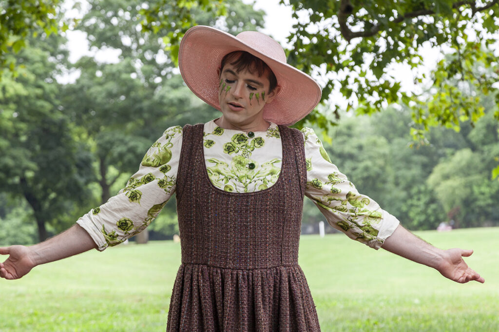Performance artist John Jarboe standing in a park with her arms stretched open wearing a pink, wide-brimmed hat and a brown jumper with a green and white patterned hand-made button down shirt underneath.