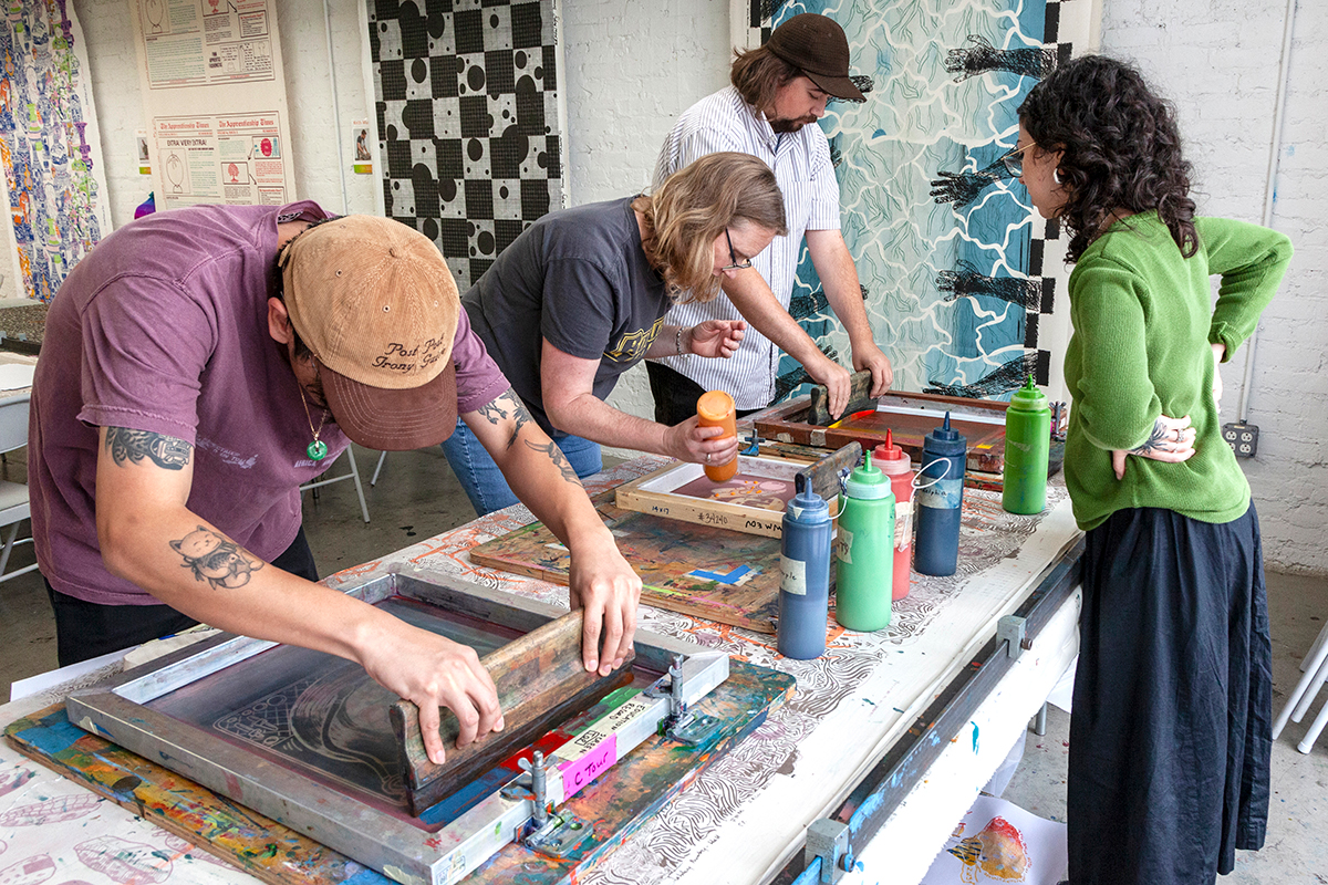 A group of three people screenprint at individual stations along a table with colorful bottles of ink nearby. A friend looks on as the group make their art.