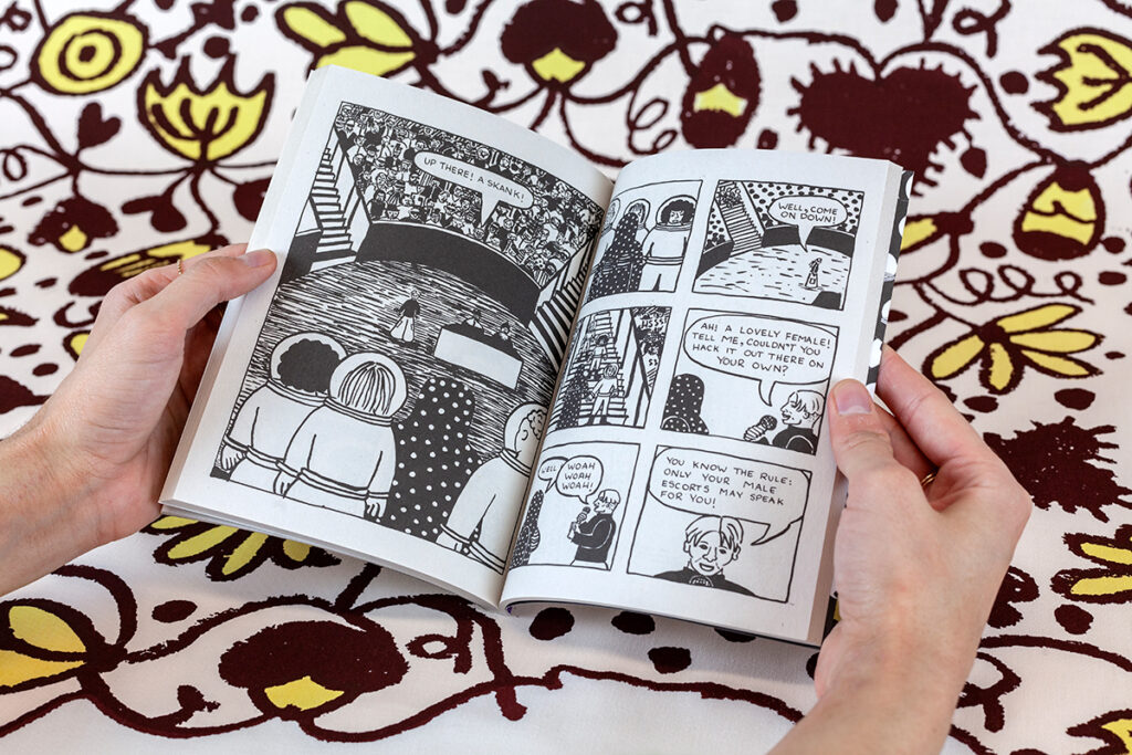 A photograph of two hands spreading a book's pages against fabric yardage consisting of brown and yellow floral motifs. The book features cartoon panels with women astronauts bringing a cloaked prisoner to a council meeting at the center of a packed legislative setting.