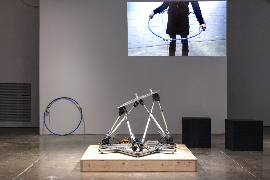 A photograph of a dimly lit gallery facing a wall with a projection of a person holding a hula hoop. The physical hula hoop leans against the wall at left while in the center foreground, a robotic armature stands atop a plinth.