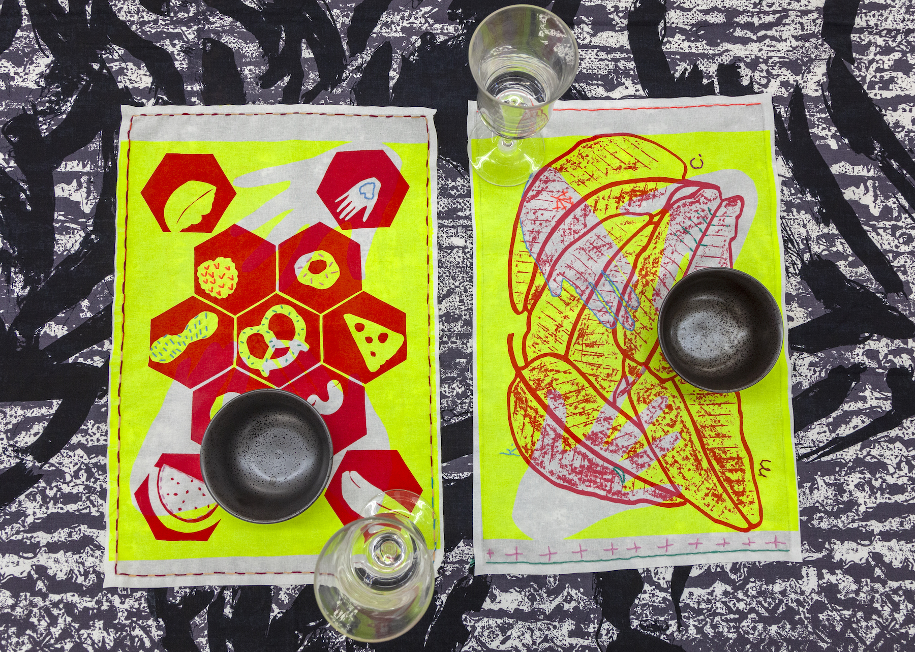 A photograph of two rectangular placemats made of fabric with a set of glasses and bowls overtop. The placemats each have a unique design printed overtop in red and bright yellow colors. On the left is a design of hexagons containing different food shapes such as pizza, pretzels, and berries. On the right is design of piled banana leaves.