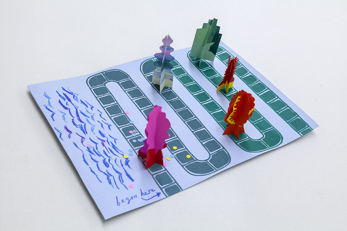 A photograph taken from above of a rectangular sheet of paper with a winding path for gameplay. On its surface are several paper game pieces that stand upright.