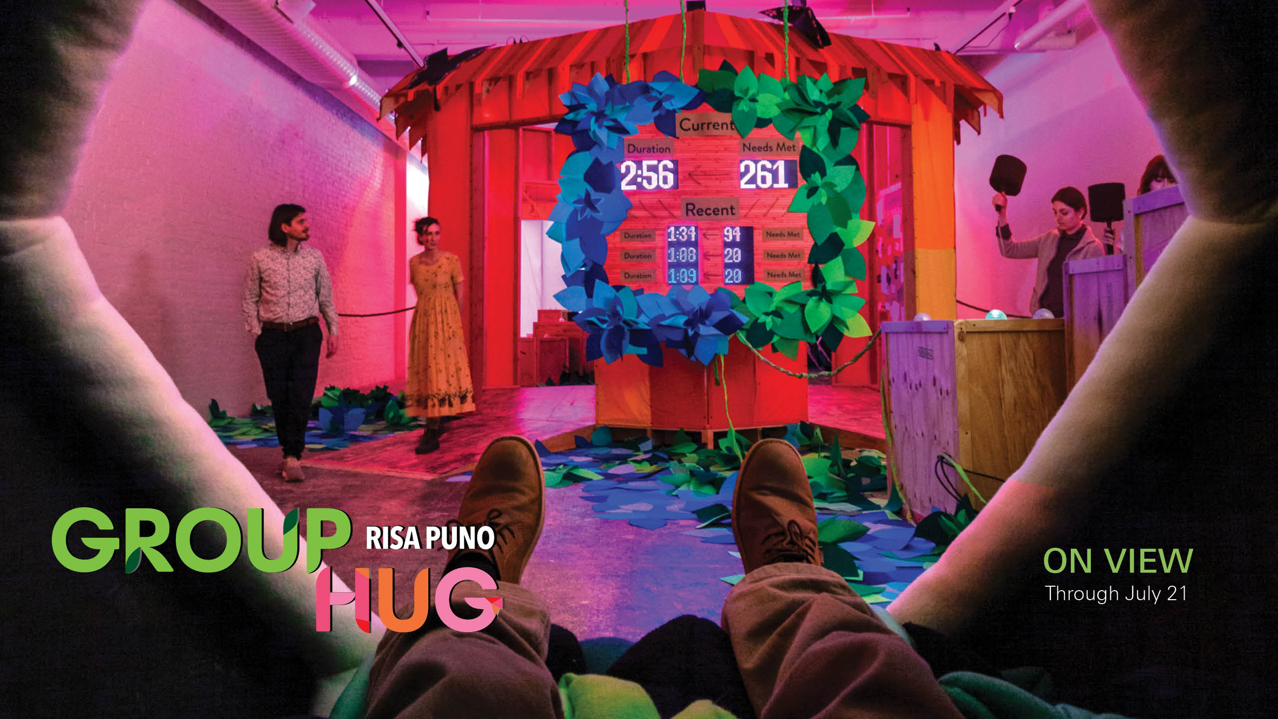 A view from inside a dark space, framed by a hexagonal shape and the photographer's own feet, into a room bathed in magenta-colored light. Straight ahead is a scoreboard with various times and scores. At the right, some people play a game with mallets. On the left, two people walk into the space. The text reads, 