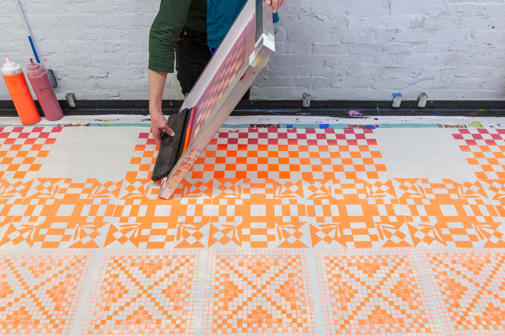 A photograph of orange geometric patterns being printed on fabric. A person lifts a silkscreen upward to reveal a newly printed design.