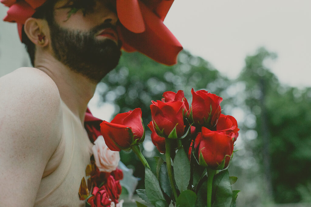 The artist John Jarboe, a white woman with a dark beard, stands outdoors wearing a red rose hat and holds a bouquet of roses against her.