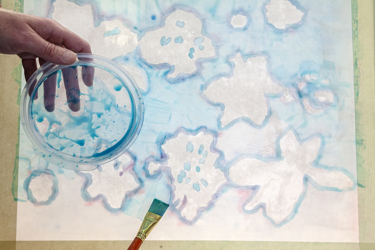 Photograph of a gestural floral painting with a light blue watery background. You can see the flat, thick paintbrush and a clear, round tupperware lid being used for a paint palette in the foreground.