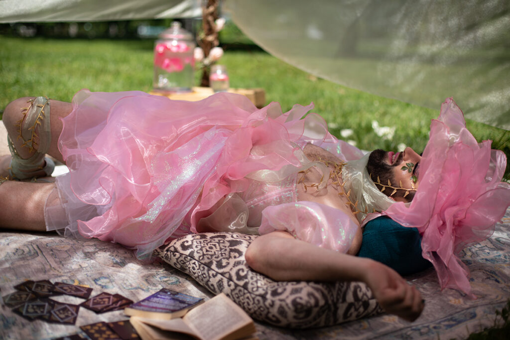 The artist John Jarboe lies down outside on a blanket and pillow, with a veiled cover shading from the summer heat. She is wearing a pink dress with a pink rose-petaled hat.