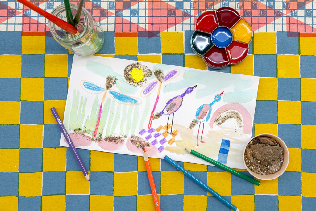 A drawing of birds and flowers using watercolor and colored pencils lies on top of a larger fabric surface that features blue and yellow checkers. A jar of brushes, a palette of ink, a cup of mica, and colored pencils lie nearby.
