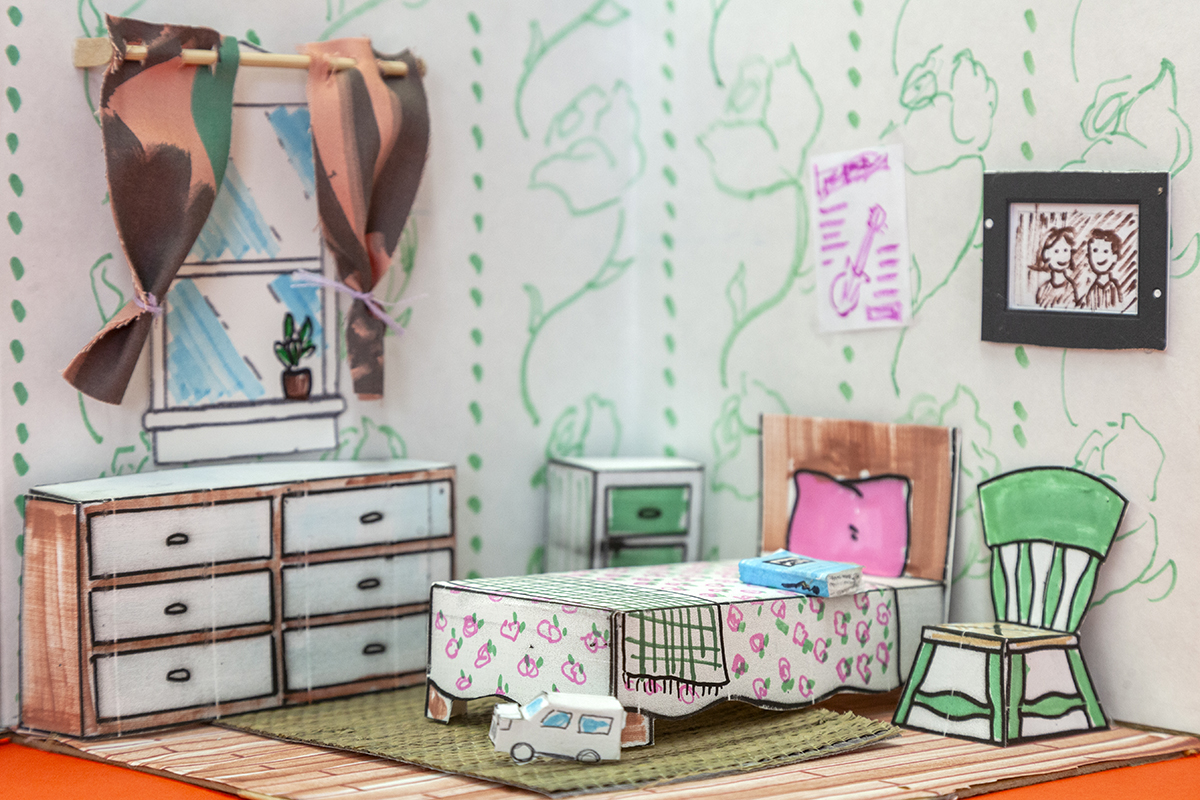 A miniature bedroom made of paper structures that are colored with patterns and pastel colors. There's a dresser, nightstand, single bed, and chair in the miniature bedroom.