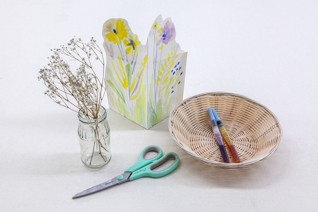 An arrangement of objects on a table, including a pair of scissors, a glass jar of wildflowers, a basket with ink tubes, and a folded sheet of paper that stands upright that features watercolor paintings of flowers.