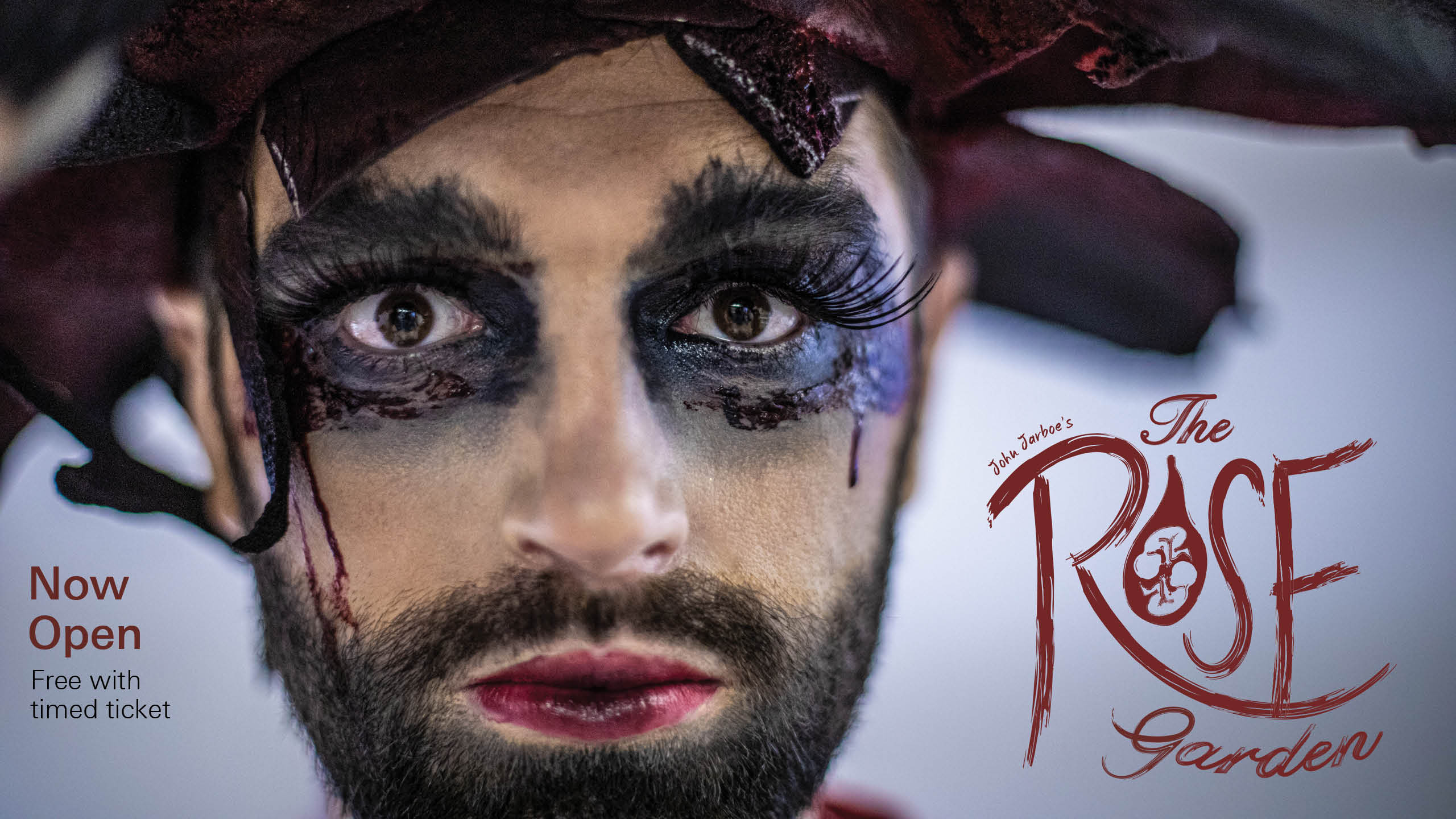 A close-up portrait of the artist John Jarboe, a white woman with a dark beard, who stares at the camera with broad dark eye make-up and a deep red wine-colored hat made of rose petal-like forms. The text reads 