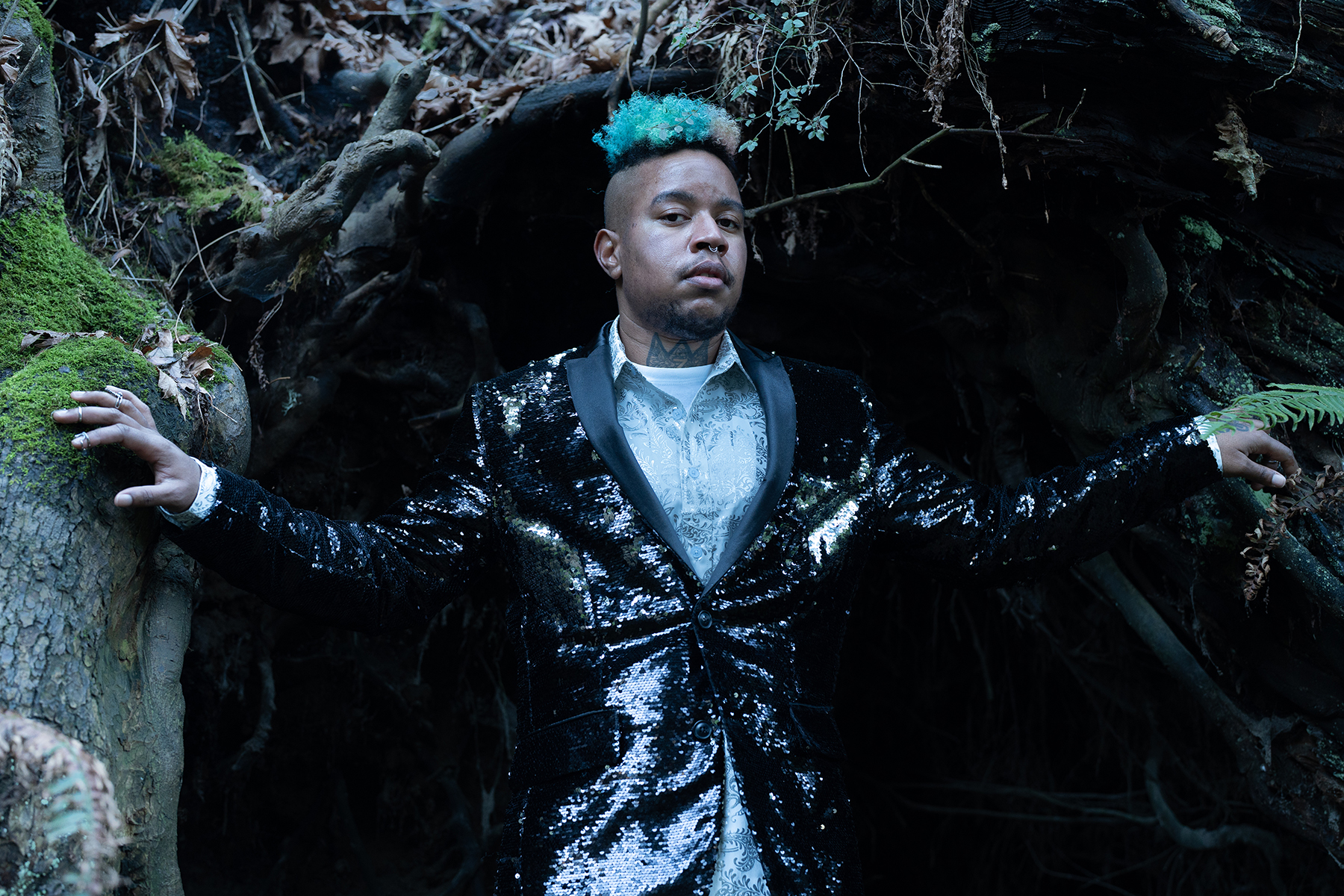 A photo of a Black man outdoors, with arms outstretched to reach each side of a cave-like hole behind him. His hair is dyed a blue-green and he is wearing a shiny black jacket.