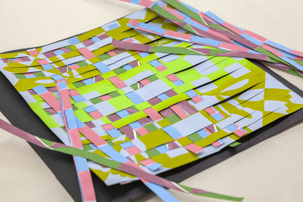 An artwork made of colorful woven paper strips. Extra paper strips lie fanned out on the corners of the artwork.