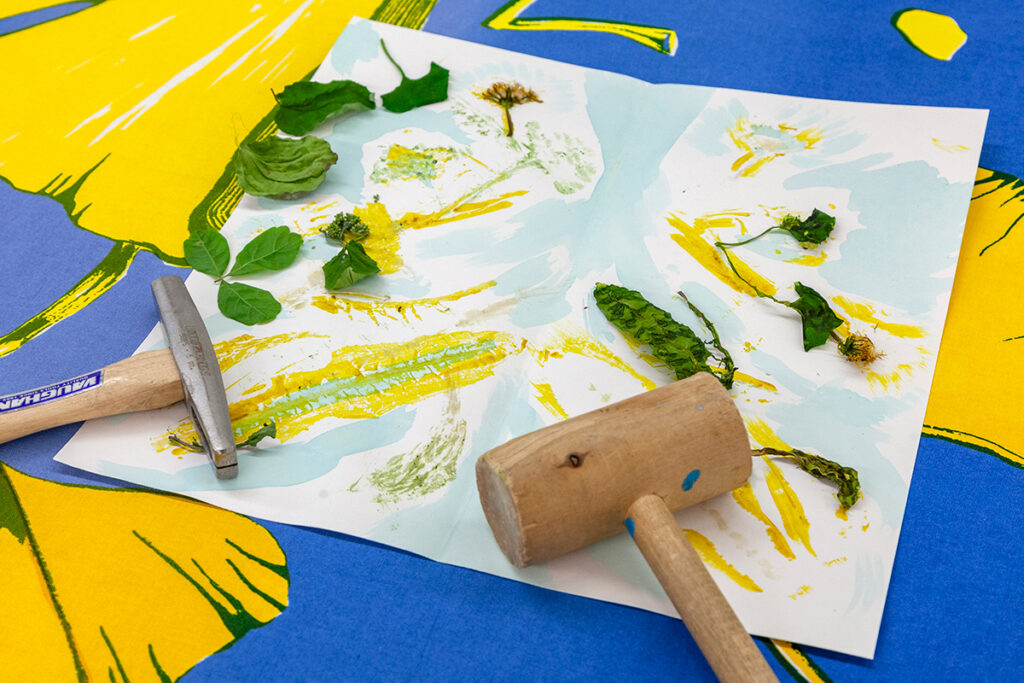 A sheet of paper with watercolor-like marks of blue and yellow lies on top of a larger blue surface with yellow leaf patterns. Various green leaves lie on top of the drawing near a mallet and tack hammer.