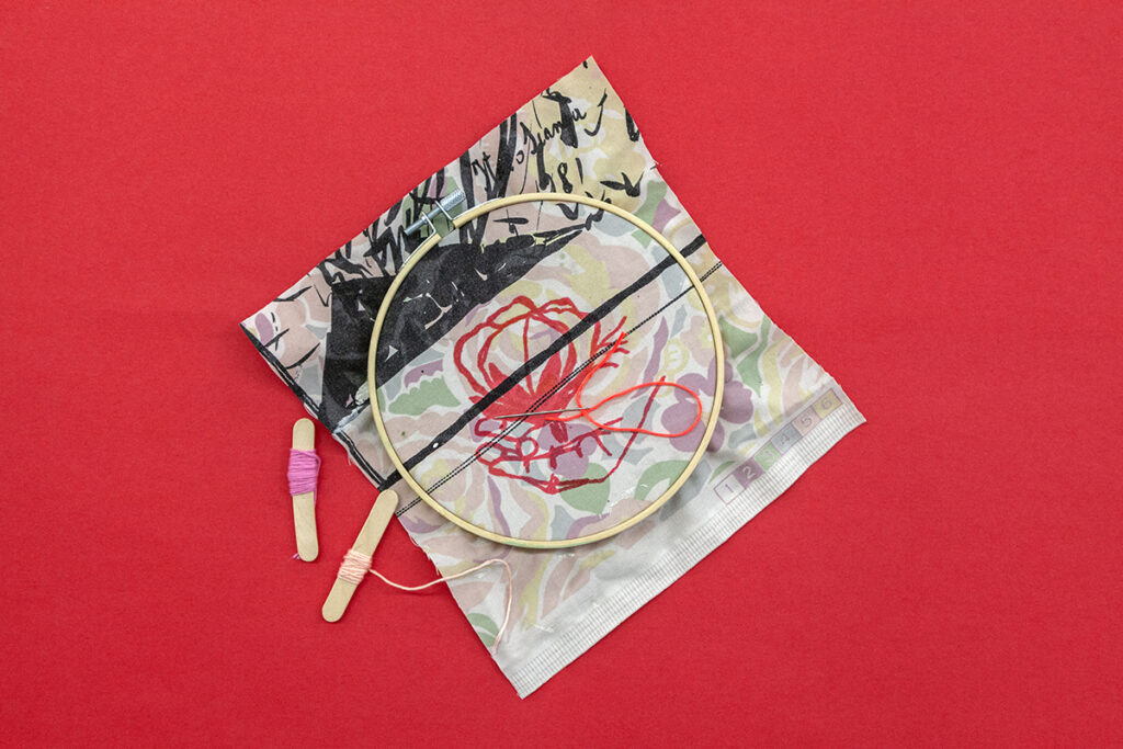 A swatch of fabric with various patterns lies on a red surface. Overtop the fabric is a wooden hoop encircling a red rose drawing.