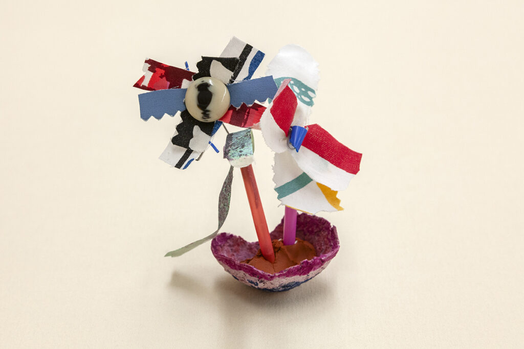 Scraps of fabric overlap one another with a button in the center, suggesting a flower. They stand on a couple of pencils planted in a small bowl of clay.
