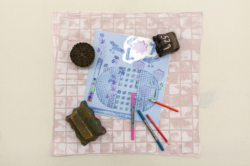 A blue square-sheet of paper lies in the middle of a soft mauve-colored set of square fabrics. The blue paper features various patterns printed from a set of etched blocks nearby. A couple colored pencils and markers lay overlapping the paper and fabric.