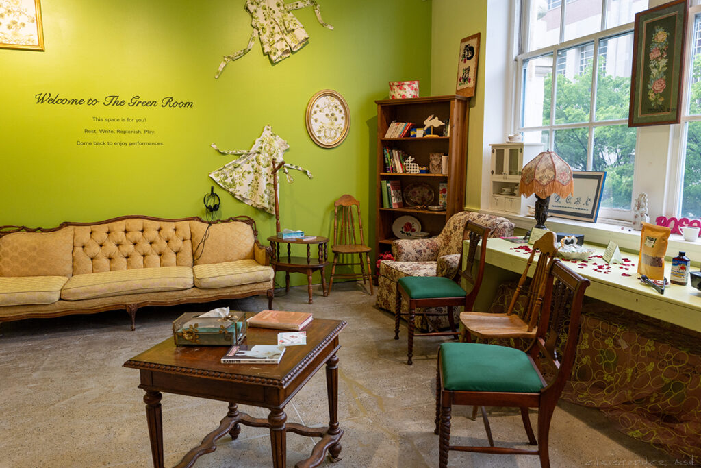 A room of vintage furniture including a table, chairs, bookshelf, and a fancy couch. The lime green wall reads "Welcome to The Green Room."