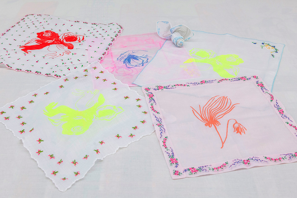 Five cream-colored handkerchiefs with lie on a table. Each features a decorative border with a floral drawing in the center, printed in different colors.