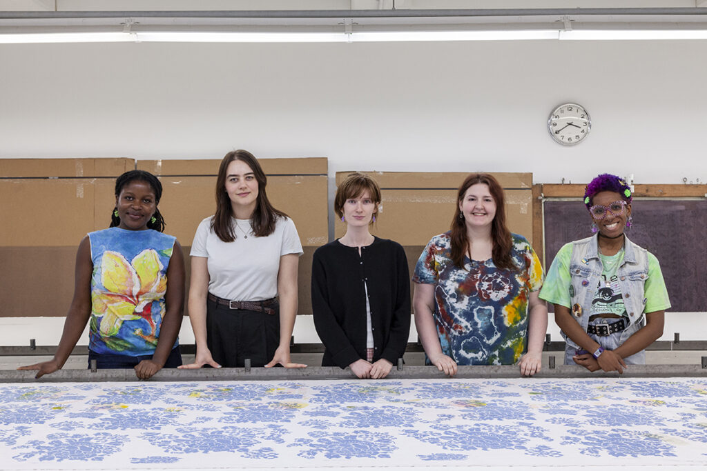 A photograph of FWM's Summer 2024 College Post Graduate Apprentices in the Print Studio. From left to right are artists Havily Nwakuche, Isa Dorvillier, Andy Marlowe, Emily Dormier, and Kalila Jones, each facing the camera.