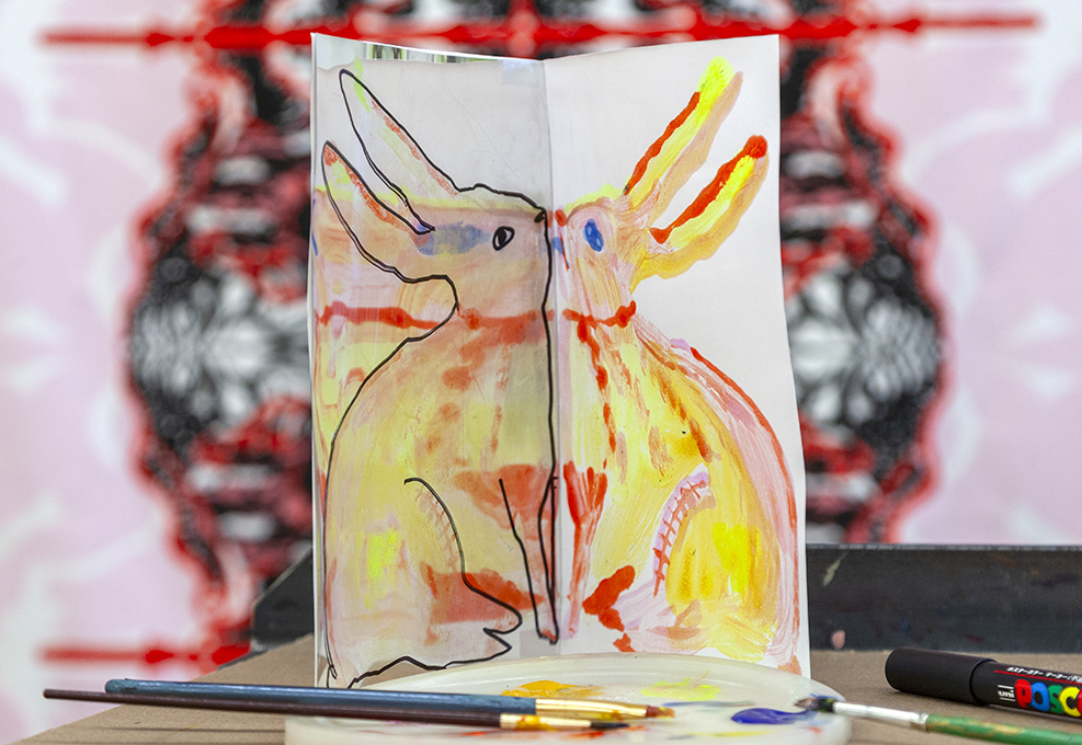 A picture of a folded sheet of paper revealing a print of a rabbit reflected on each side, their faces touching at the centerfold. Watercolor brushes lie nearby.