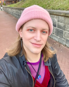 A portrait of Pax Ressler, a white non-binary transfeminine person with should-length blonde hair. Standing outdoors, they are wearing a pink beanie, black jacket, and purple-colored shirt.