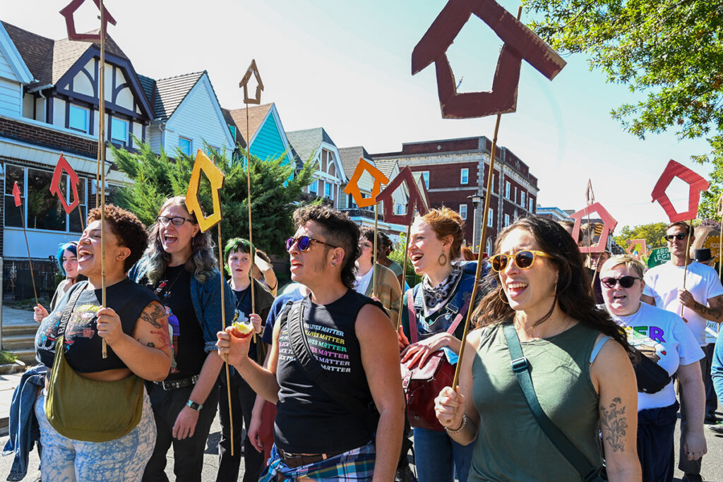 A photo of a group of people marching through a neighborhood. They are holding rods with house-shaped symbols, made in different colors.