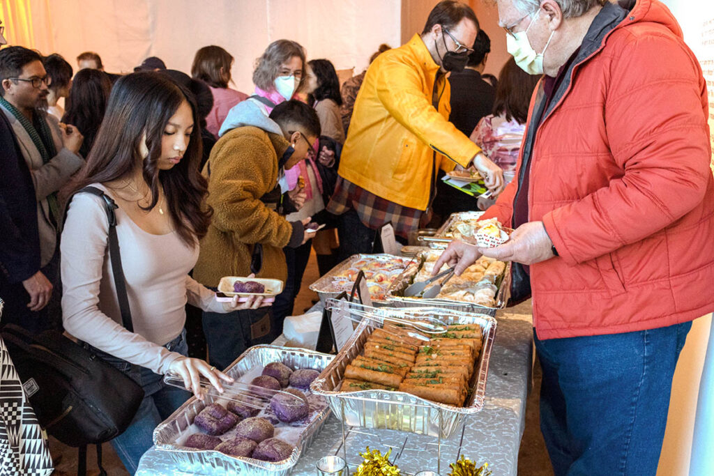 A group of people gather around a table filled with Filipino food in serving trays. At left, a young woman draws from a tray of purple breads. At right, a man uses tongs to pick up some fried rolls.