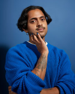 A portrait of Krishna Istha, a comedian of South Asian descent, who looks at the viewer with their right hand pressed against their jaw. They are wearing blue nail polish and a blue sweater with sleeves pulled back to reveal a flora-inspired tattoo on their right arm, while standing in front of a blue backdrop. They have a light mustache and wavy black hair, which rests at their ear.