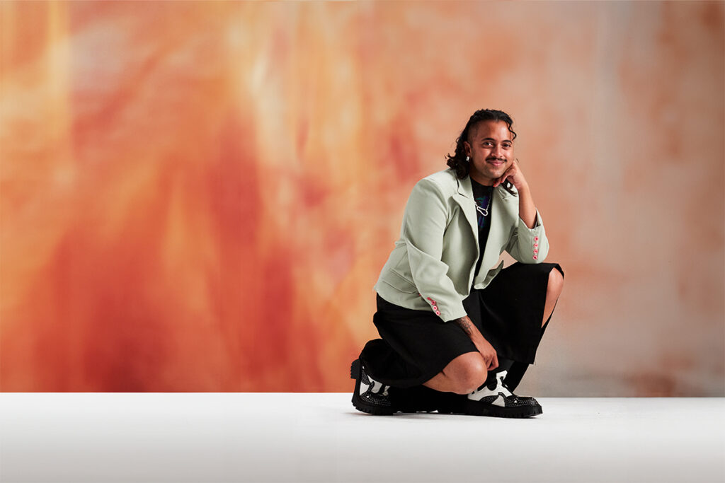 A portrait of Krishna Istha, a comedian of South Asian descent, who is posing for the camera in a crouched position. Their face rests on their left hand; their elbow propped up by their knee. They are wearing a light olive blazer, a loose black skirt, and high top sneakers. Behind them is a warm colored back-drop.