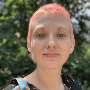 A portrait of the trans theater artist MK Tuomanen, a white person with light eyes and short pink-dyed hair. They are outdoors and smiling slightly while looking at the camera.