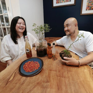 A portrait of a Filipino couple, Nicky Uy and Omar Buenaventura, seated at a table in their home. Nicky (at left) laughs as Omar smiles at her. On the table are a couple native plants, bottles, and a plate of red seeds.